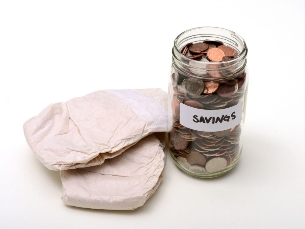 Glass jar filled with assorted coins, labeled 'SAVINGS', placed next to two folded diapers.