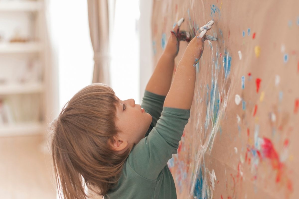 A young child in a green shirt reaching up to draw colorful pictures on a wall with paint.