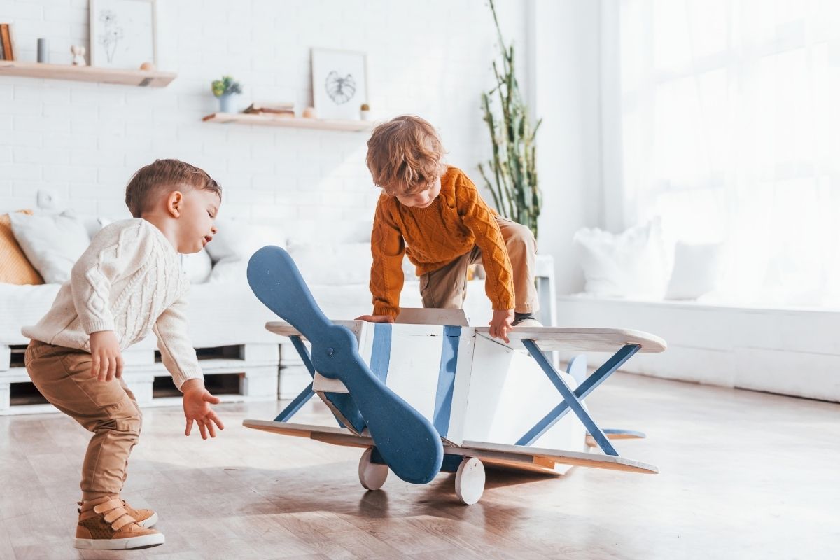 Two young children playing with a blue wooden toy airplane in a bright room.