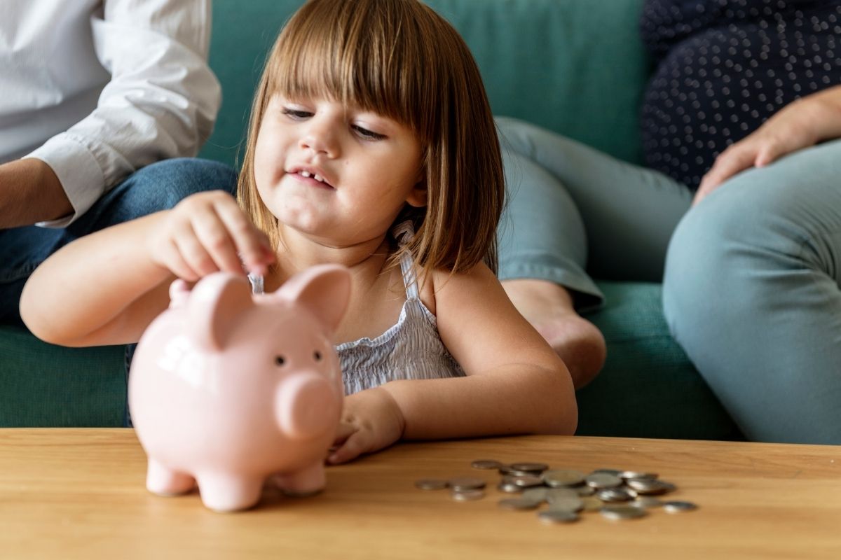 Young child putting a coin into a pink piggy bank while sitting at a table.