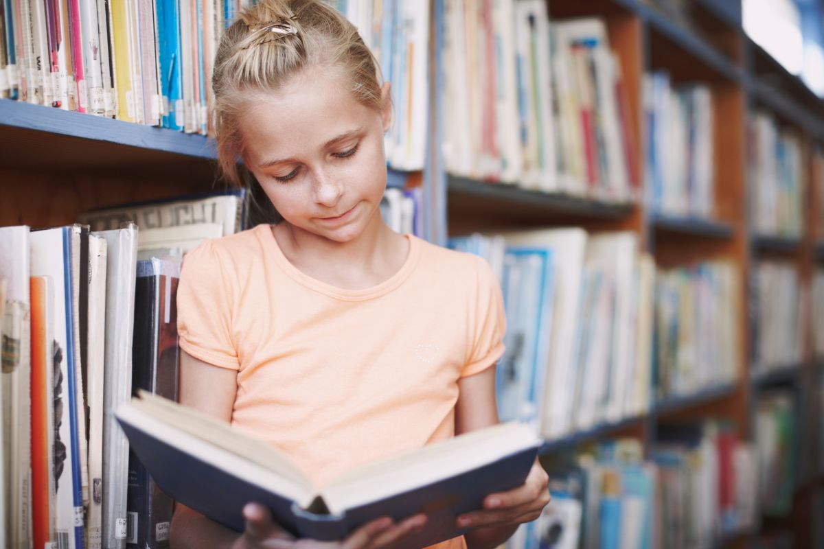 Young girl reading a book while standing between shelves in a library.