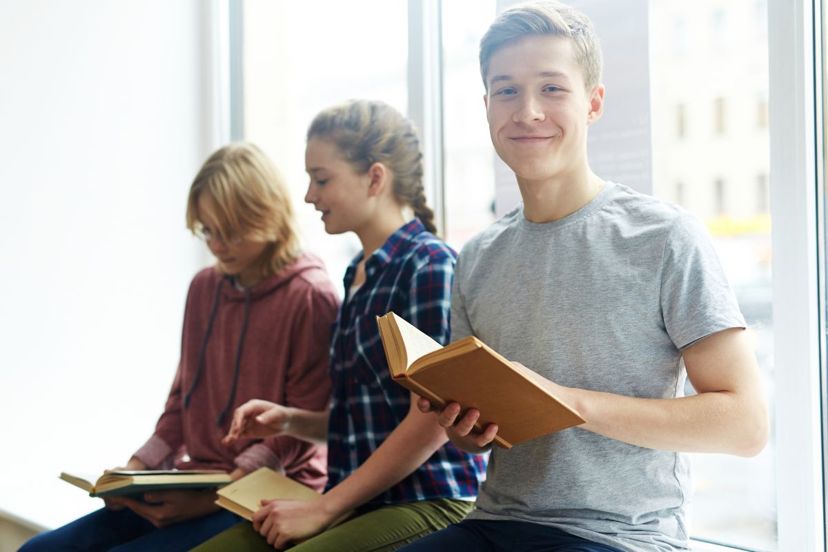 Group of teenagers reading books together, with one boy holding an open book in the foreground.