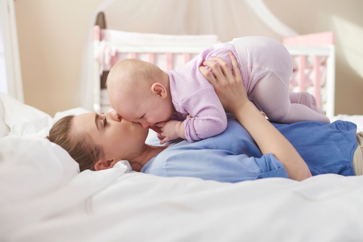 A mother lying on a bed and gently kissing her baby while holding the child above her chest.