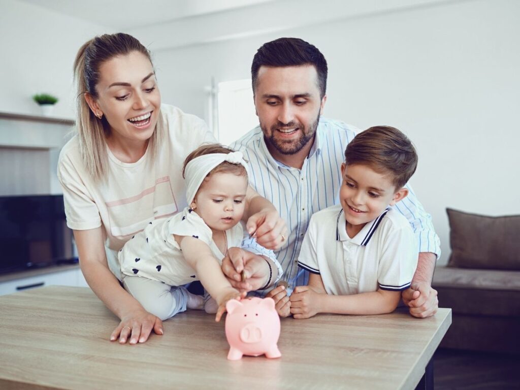 Family gathered around a table, engaging with a pink piggy bank, the mother and father putting coins into it