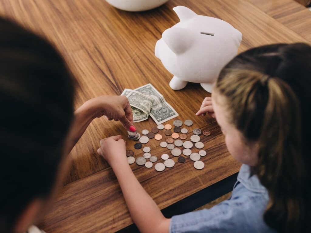 mother and daughter sitting at a wooden table, counting and organizing coins and dollar bills, with a white piggy bank nearby