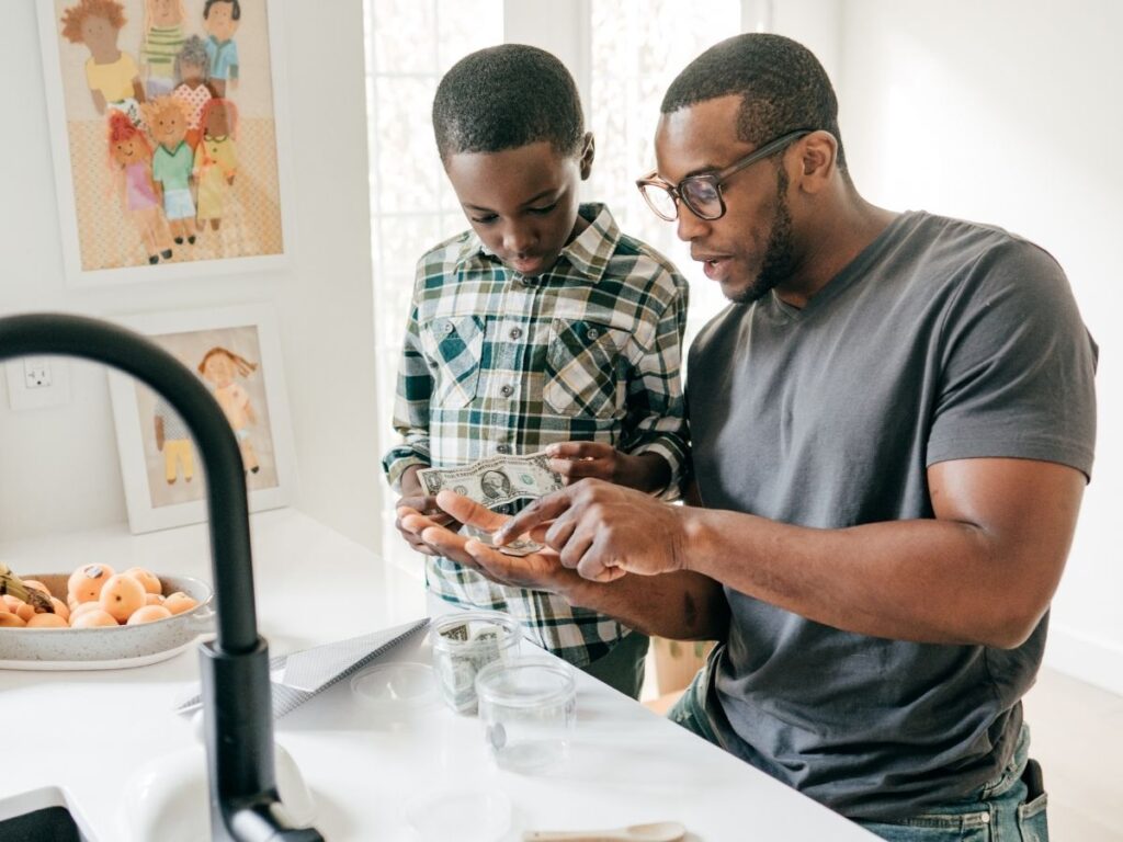 father and son at the kitchen counter holding dollar bills and coins as the father explains something to his son