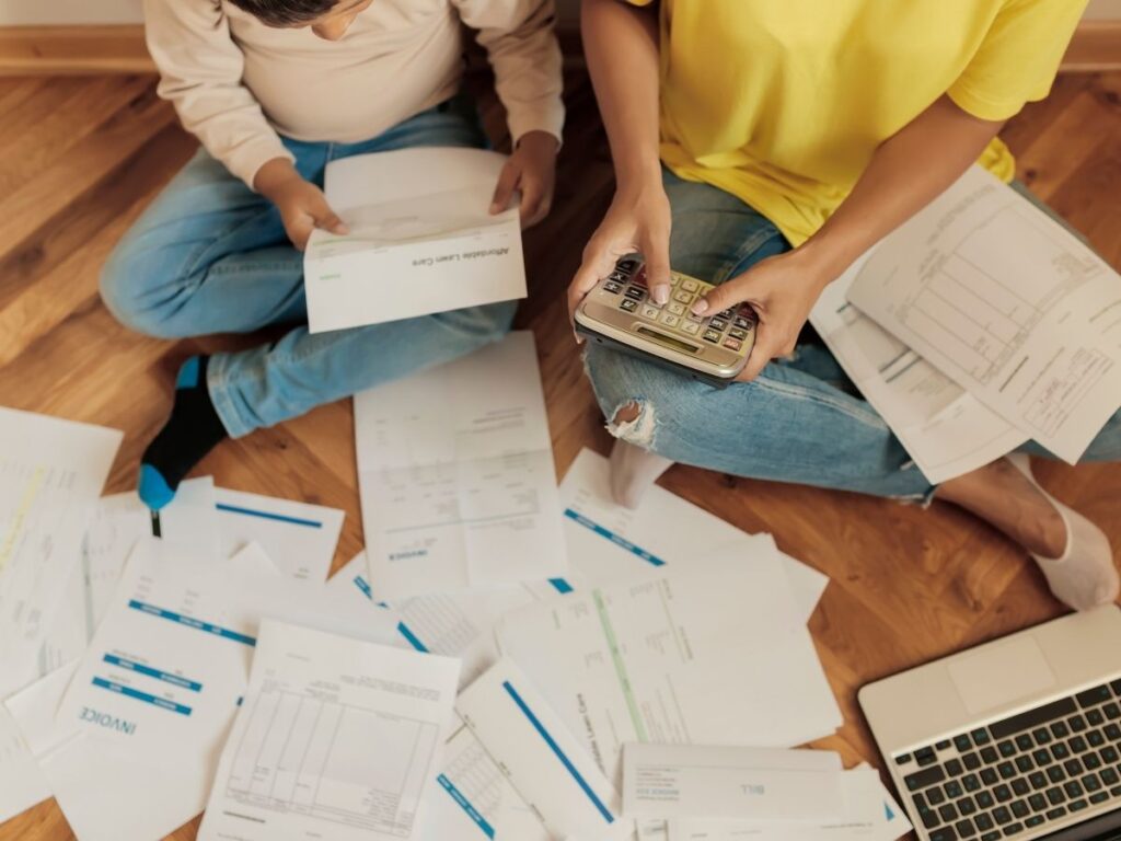 mother and son sitting on the floor surrounded by various financial documents, including invoices and bills, with the mother holding a calculator, while the kid is reviewing one of the documents