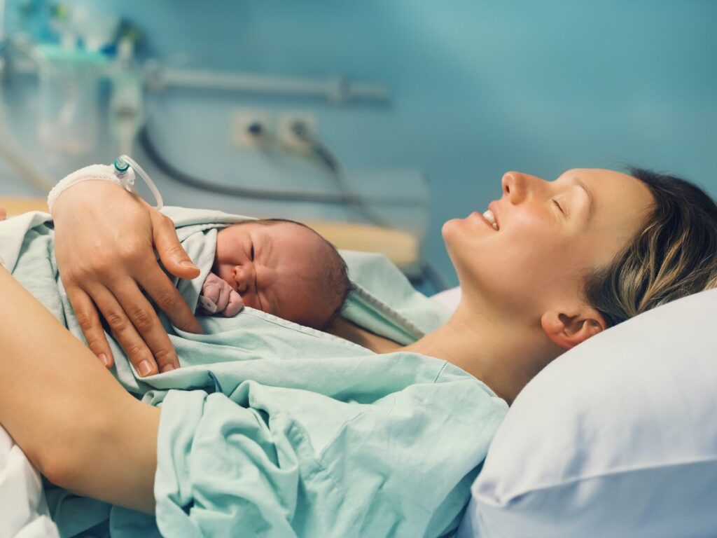Mother after birth in a hospital gown on a bed, hugging her newborn baby laying on her chest.