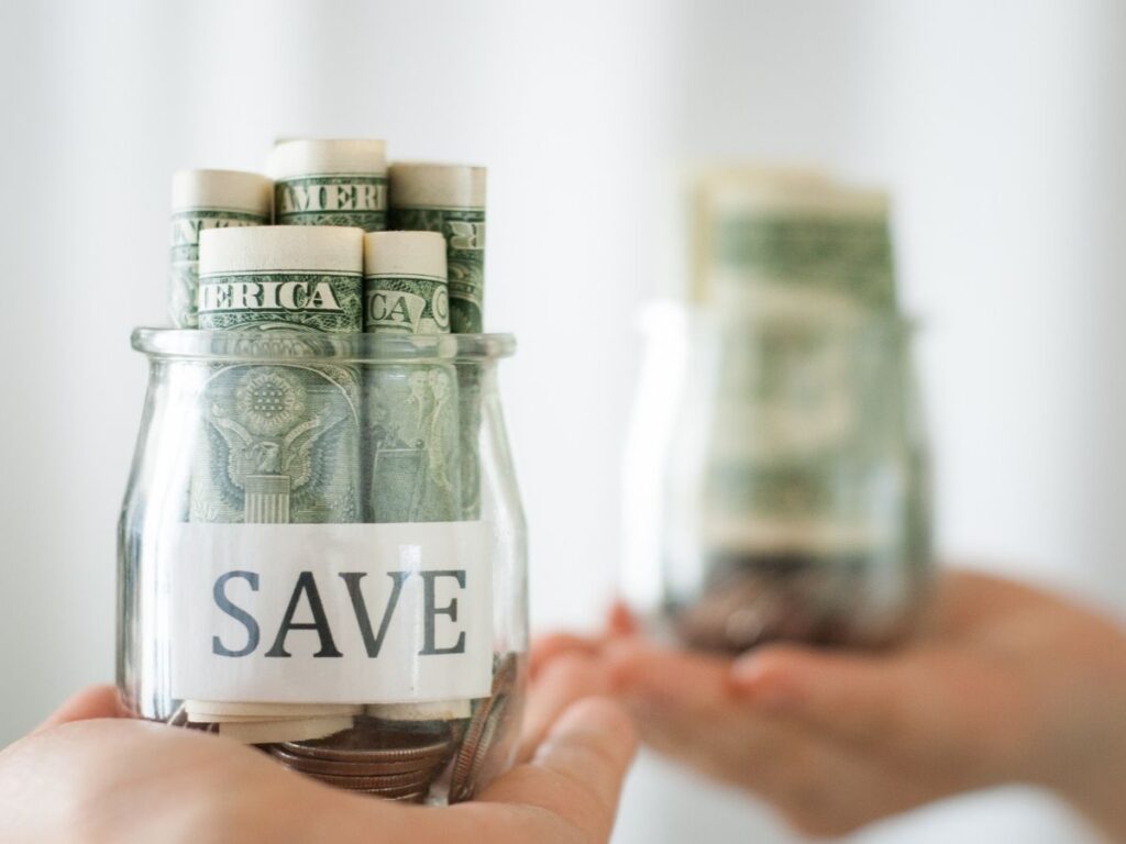Two hands holding glass jars filled with rolled-up U.S. dollar bills and coins, one labeled 'SAVE'