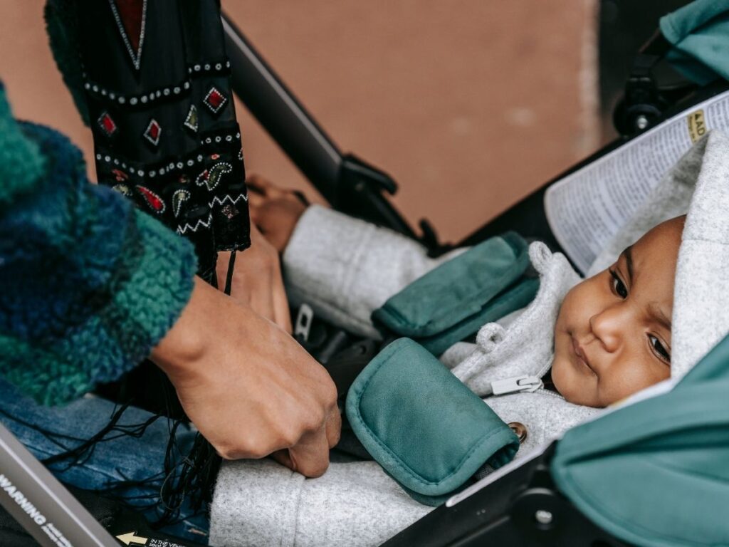 A person adjusting the straps on a stroller with a toddler wearing a gray jacket on a green padded seat