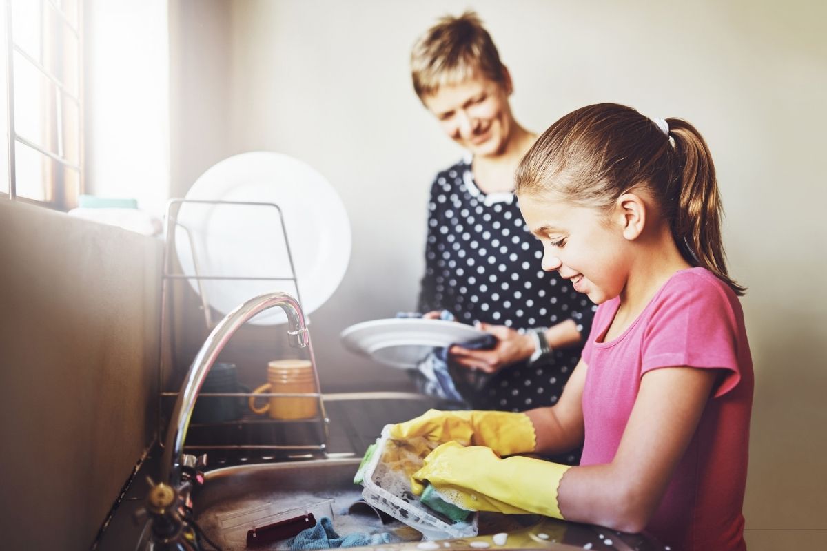 Young girl washing dishes at the kitchen sink while an adult stands beside her.