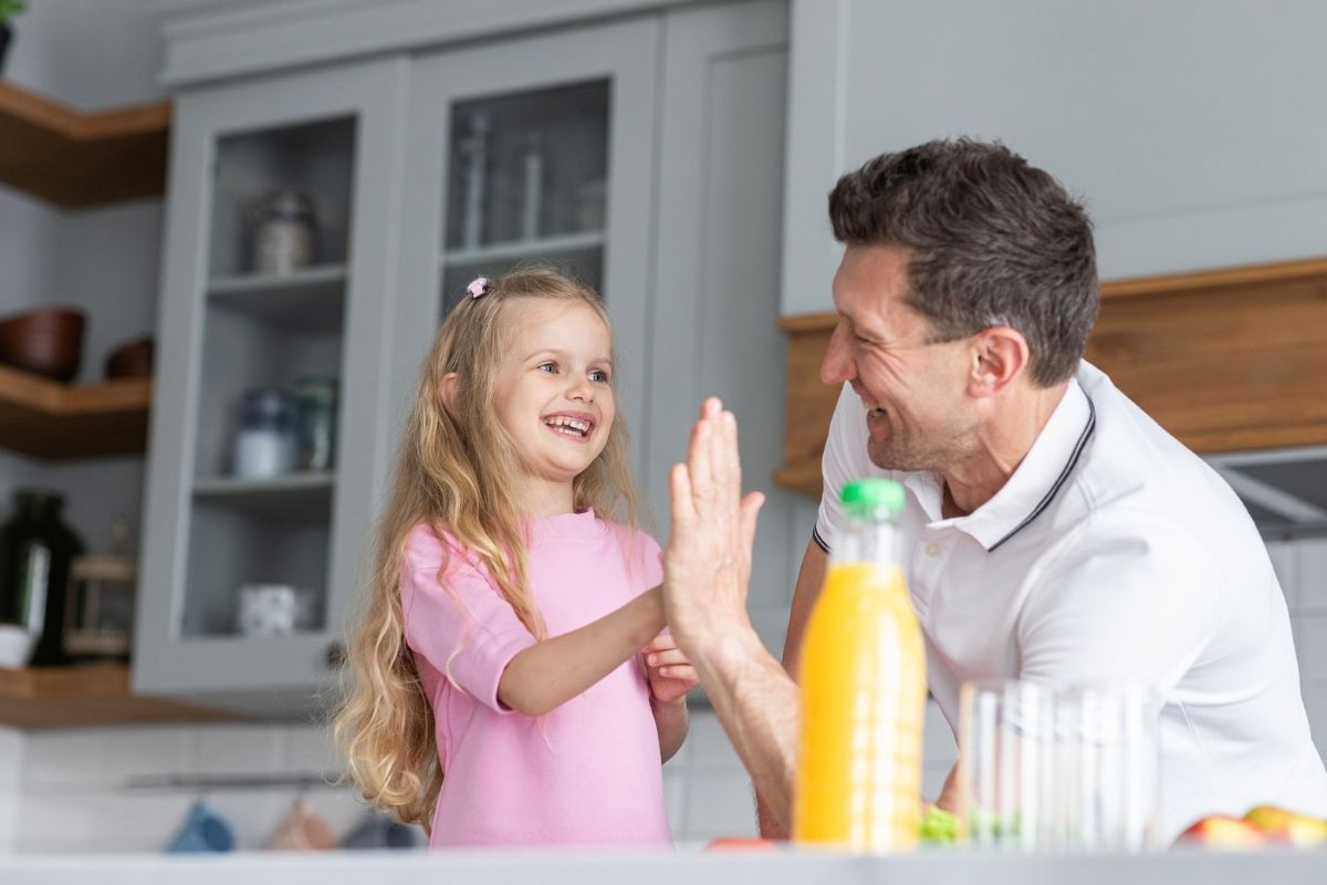 Father and young daughter giving each other a high five while doing chores together in the kitchen.