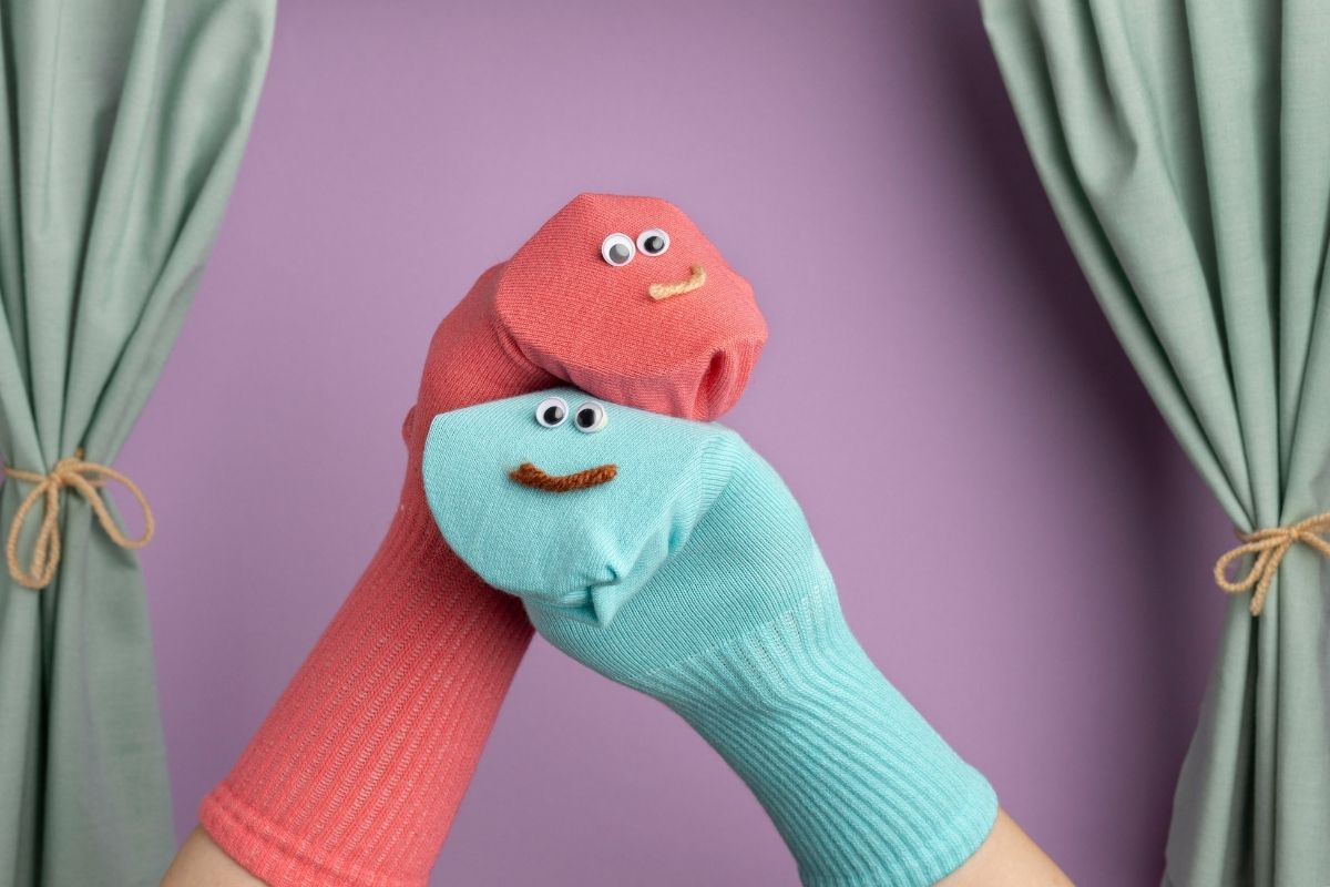 Colorful sock puppets with googly eyes performing against a purple background with green curtains.