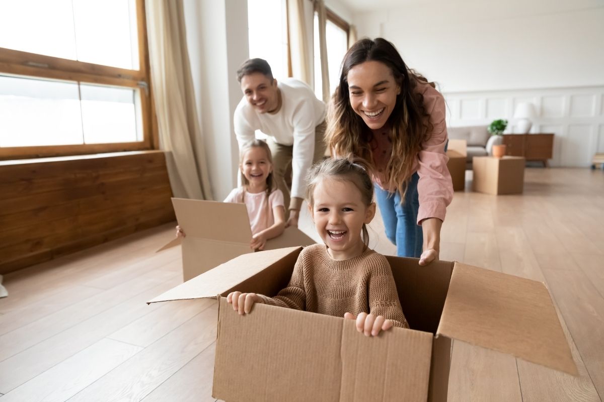 Parents playing with their two daughters in cardboard boxes on the living room floor.