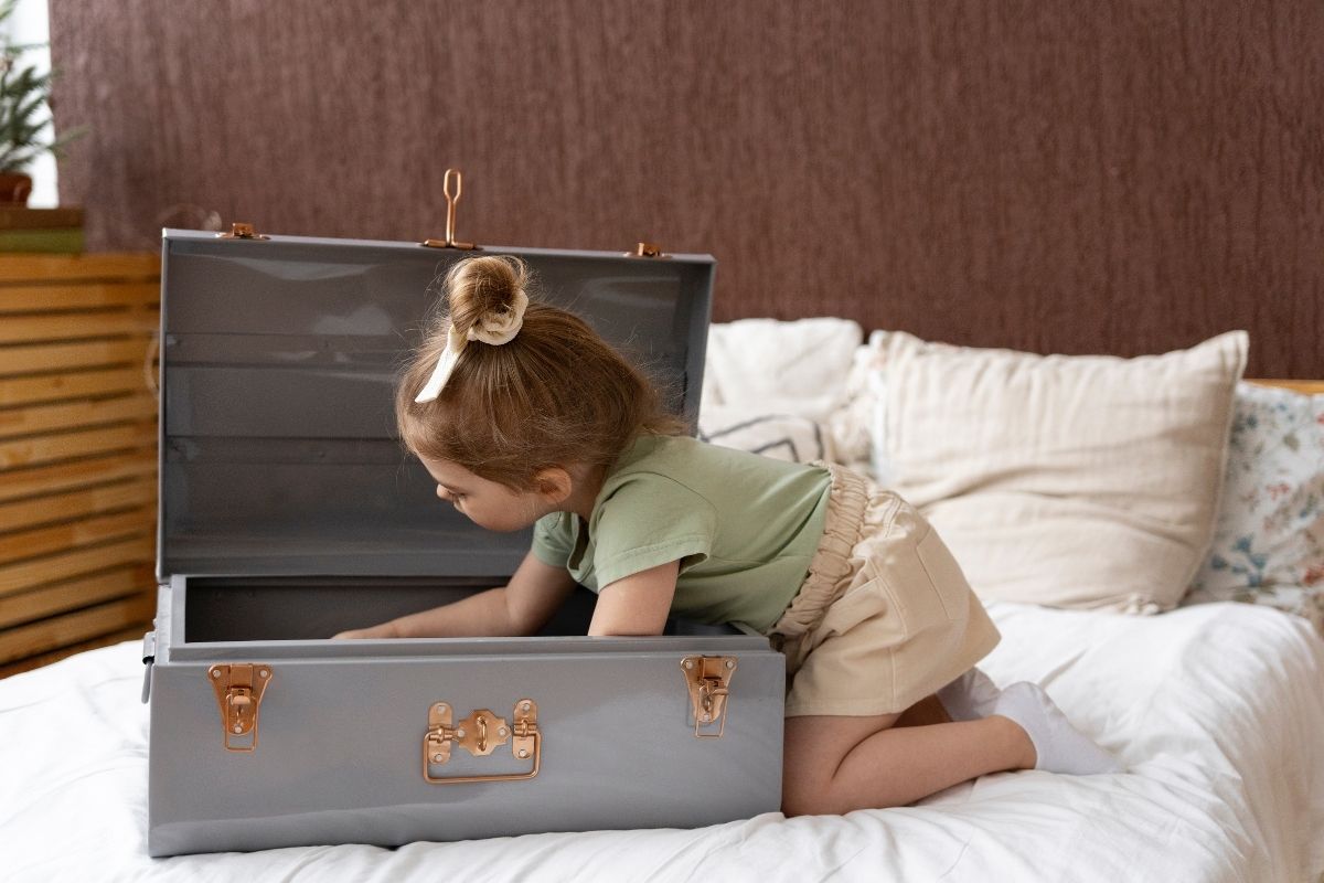 Little girl exploring an open vintage suitcase while kneeling on a bed.