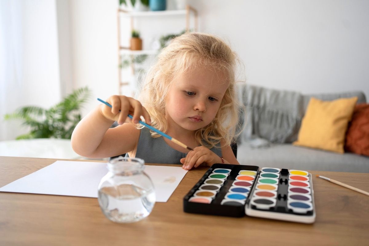 Young girl painting with watercolors at a wooden table, using a paintbrush and colorful paint set.