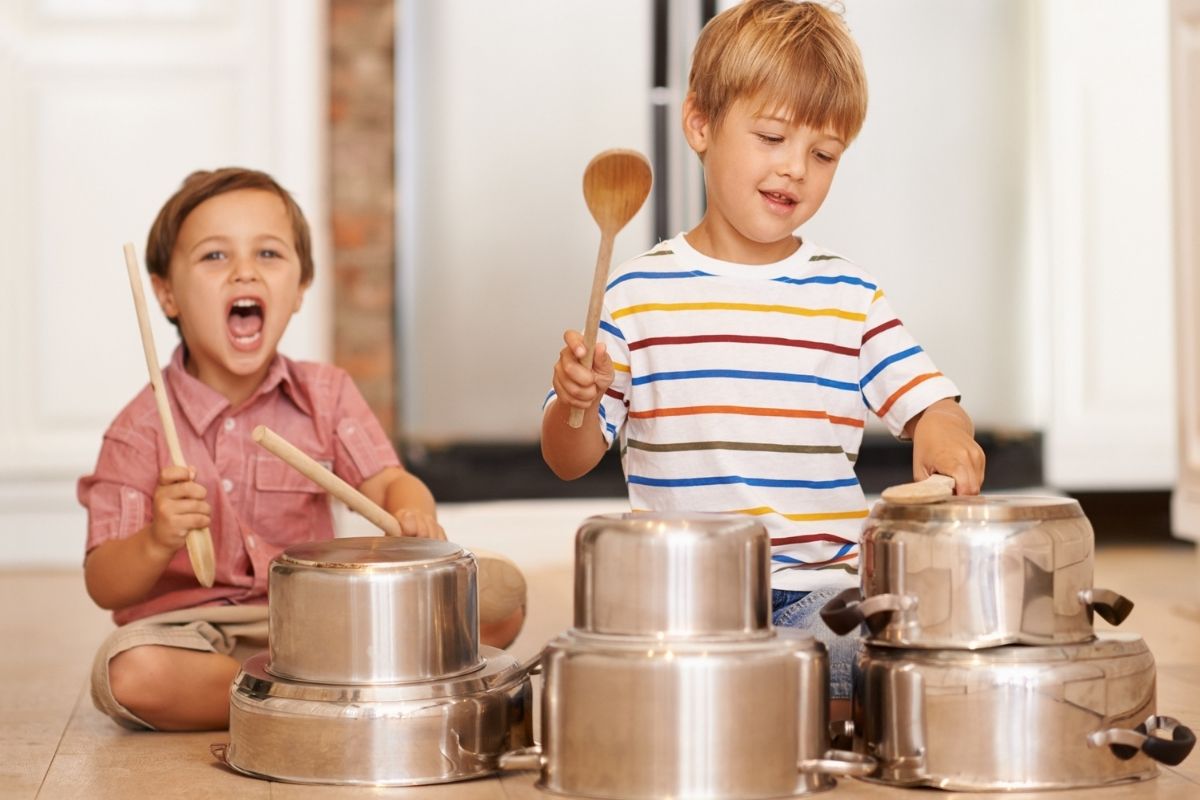 Two young children playing homemade drums using metal pots and wooden spoons on the kitchen floor.