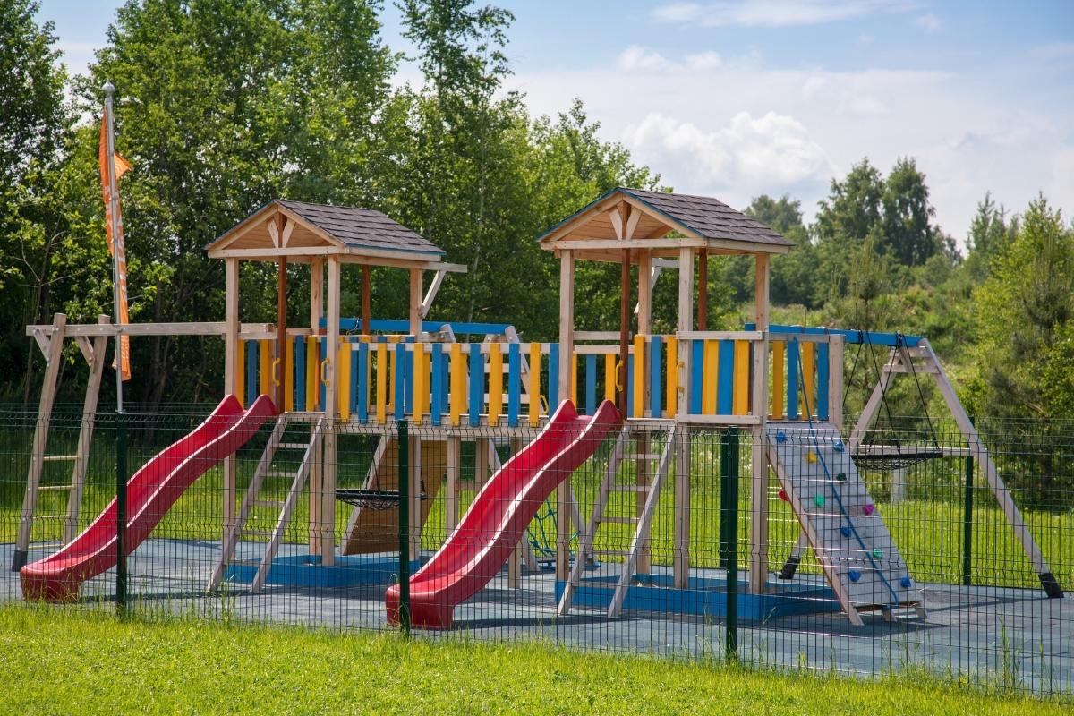 Wooden playground with two red slides, climbing ladders, and swings surrounded by green trees.