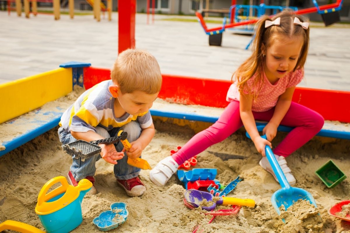 Two young children playing with colorful sand toys in an outdoor sandbox.