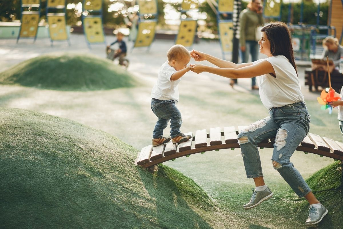 Mother helping her toddler balance on a small wooden bridge at a playground.