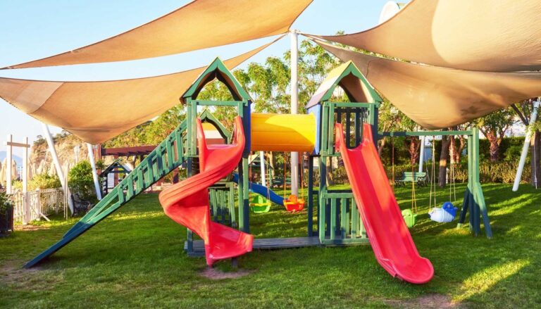Outdoor playground with red slides, climbing structures, swings, and large shade sails providing sun protection.