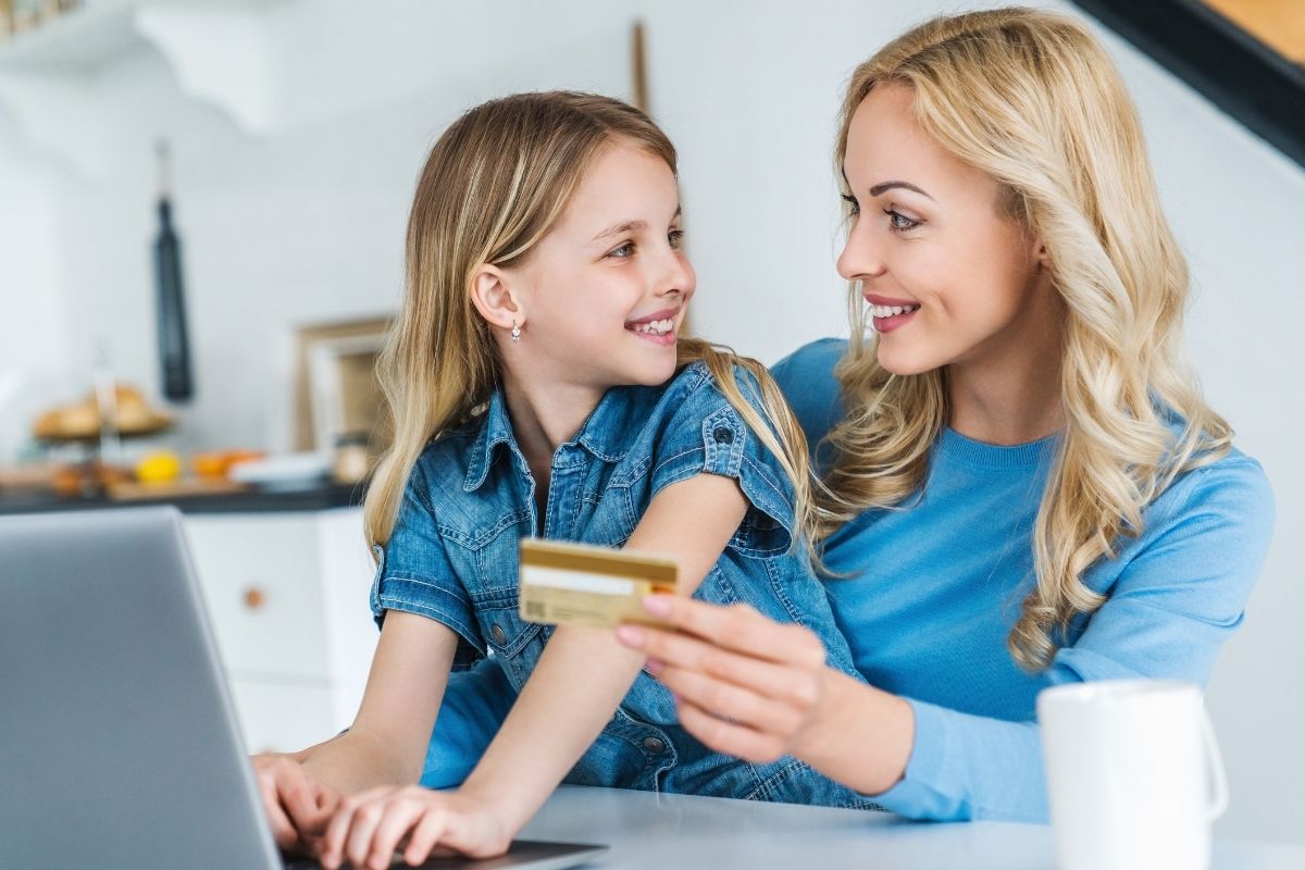 Mother and daughter using a laptop together while holding a bank card at home.
