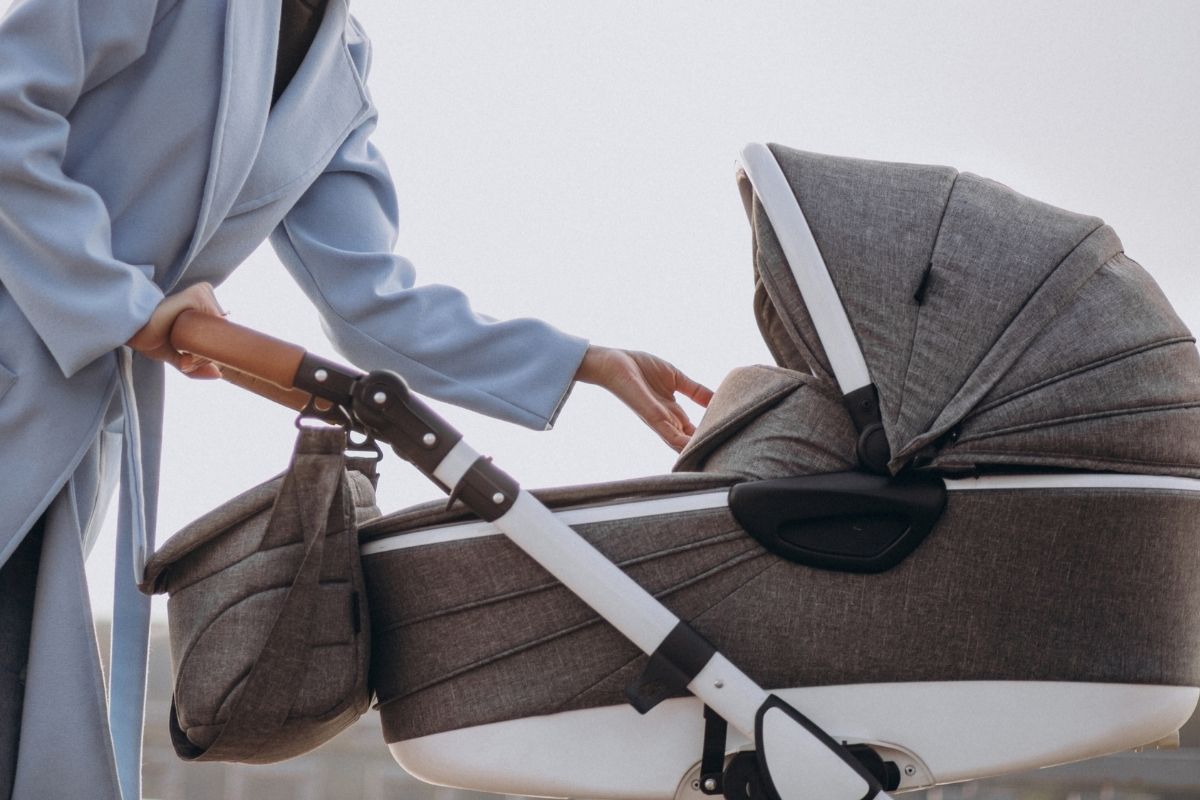 A parent in a light blue coat adjusting the canopy of a gray baby stroller with a bassinet.