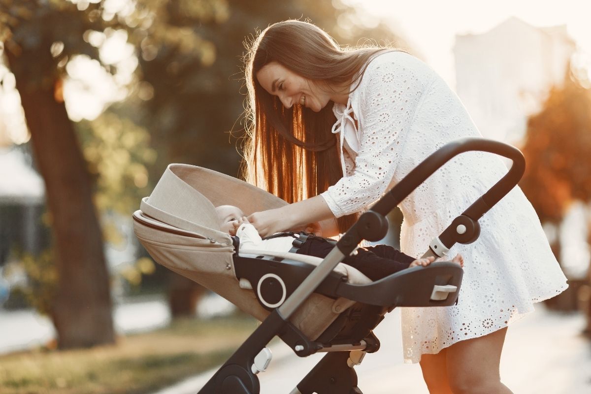 A mother in a white dress leaning over a baby stroller and checking on her child during a walk in a sunlit park.