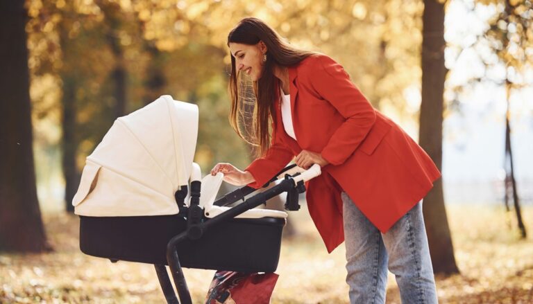 A mother in a red coat checking a baby stroller with a white canopy during a walk in an autumn park.
