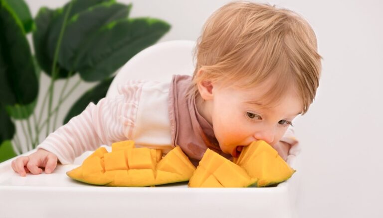 Toddler leaning forward to bite into a sliced mango on a high chair tray.