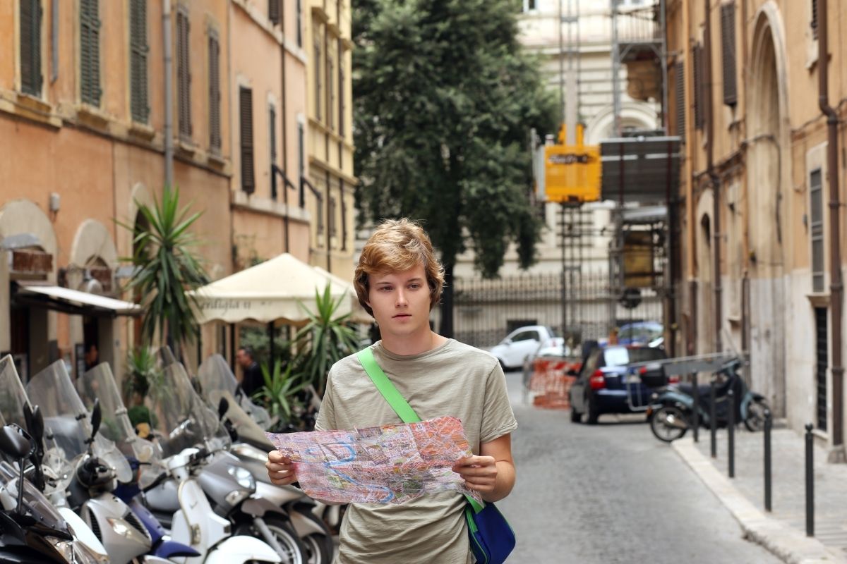 Teenage boy holding a city map while exploring a European street lined with parked scooters and historic buildings.
