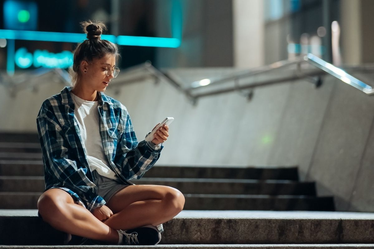 Teenage girl wearing glasses and a plaid shirt sitting on outdoor steps at night while checking her smartphone.