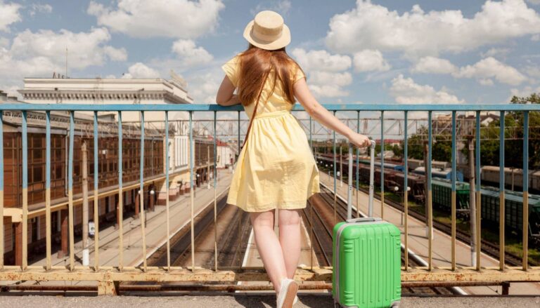 Young woman in a yellow dress and straw hat standing beside a green suitcase on a pedestrian bridge overlooking railway tracks.