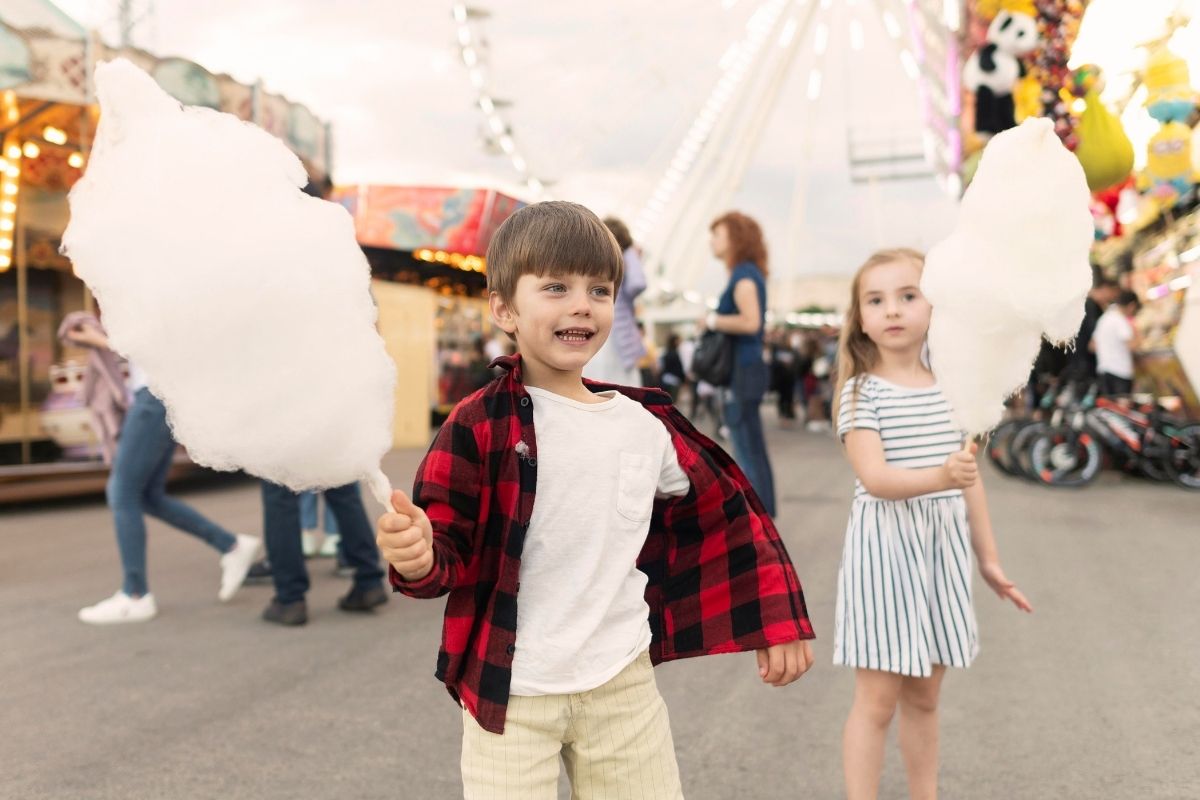 Two children holding large sticks of cotton candy while exploring a lively amusement park with rides in the background.