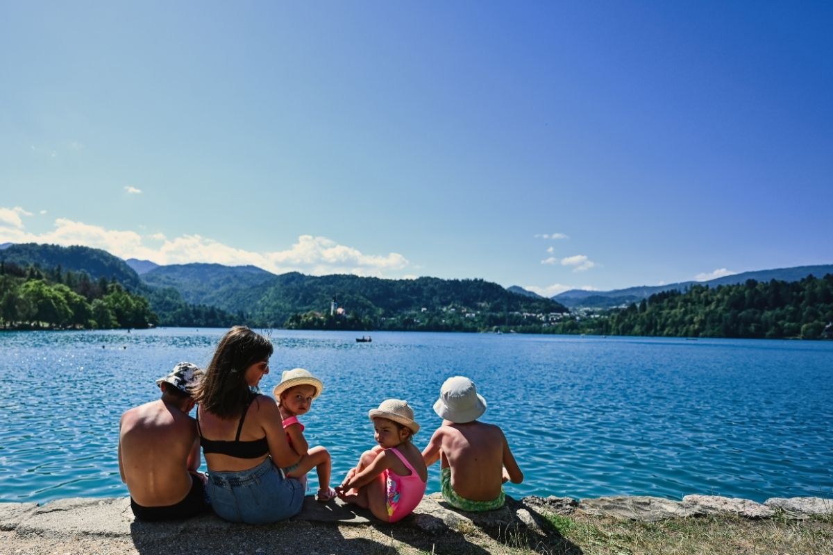Family with two young children sitting on a rocky lakeshore, enjoying a scenic view of the water and surrounding mountains.