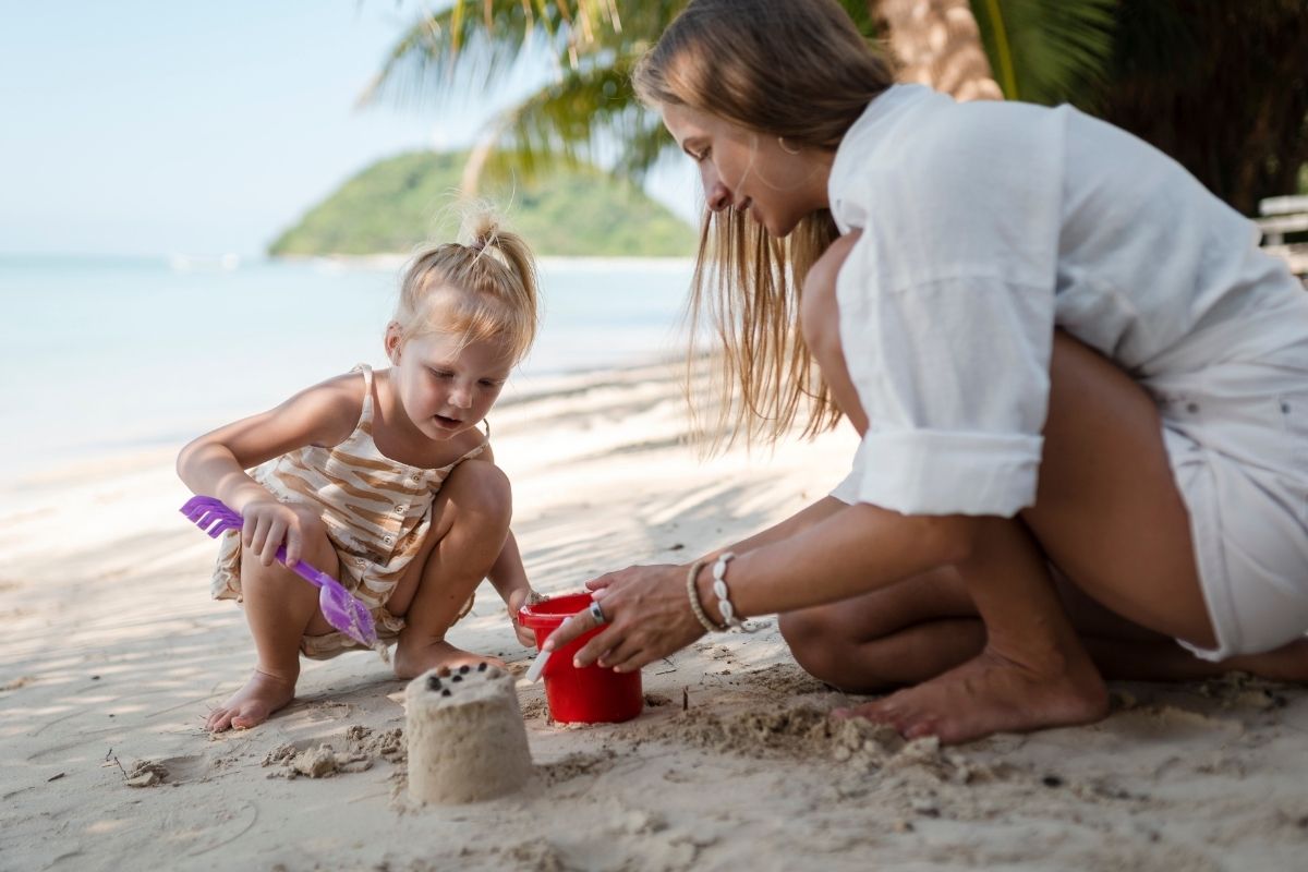 Mother and young daughter building sandcastles together on a tropical beach with palm trees and the sea in the background.