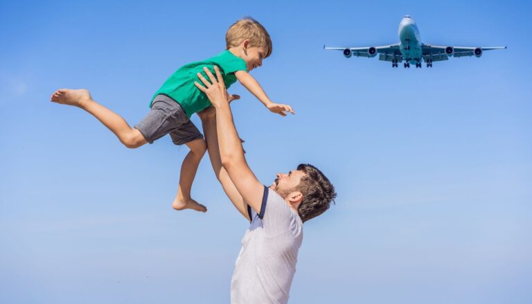 Father lifting his young son into the air outdoors, with an airplane flying overhead against a clear blue sky.