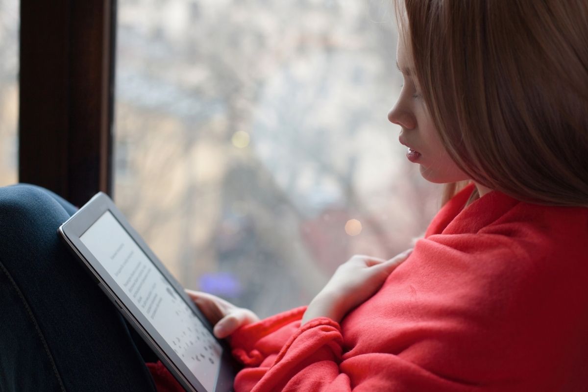 Teenage girl in a red sweatshirt reading an e-book on an e-reader beside a window.