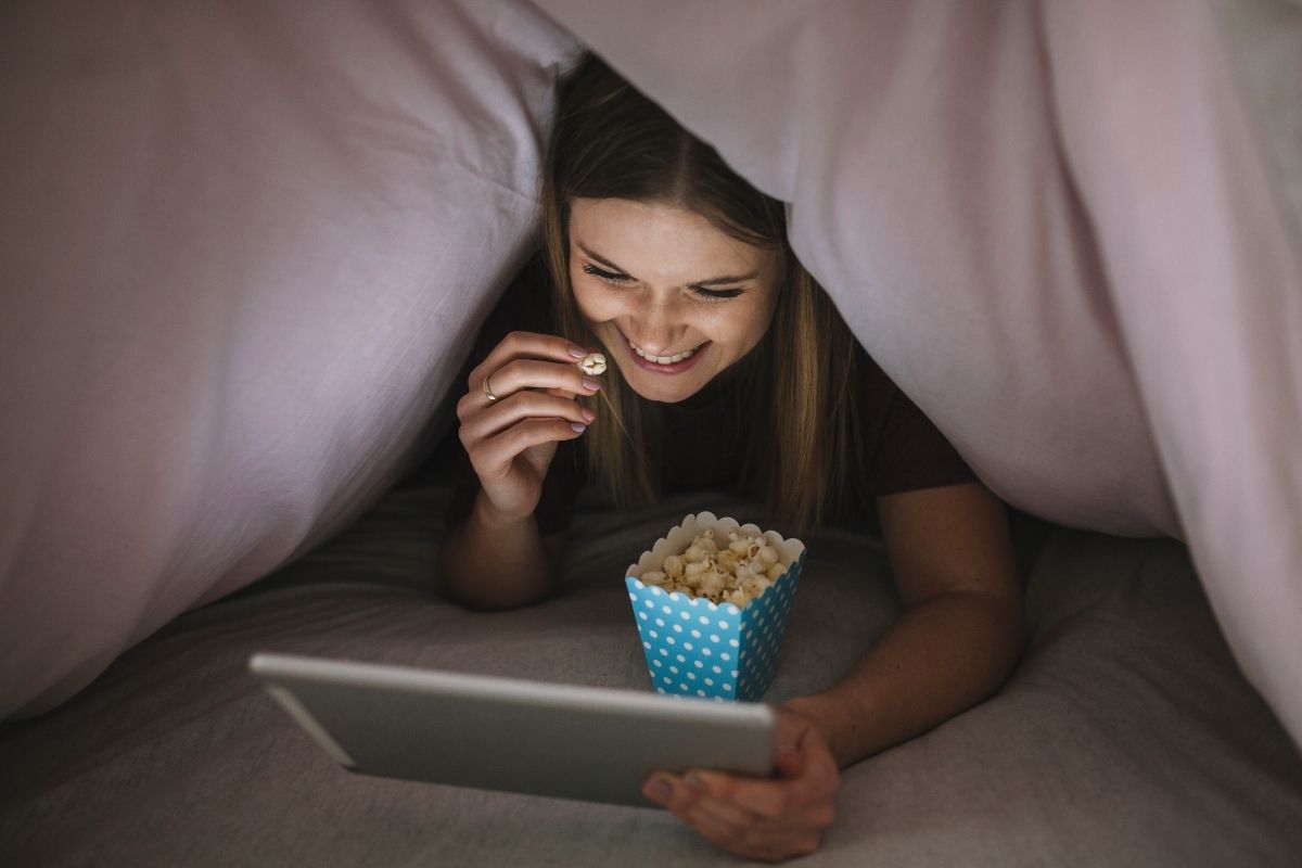 Smiling teenage girl watching a tablet and eating popcorn under a blanket.