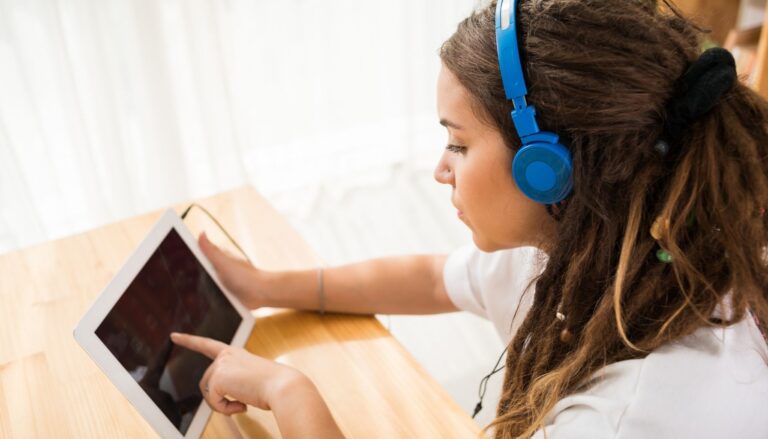 Teenage girl wearing blue headphones and using a tablet while sitting at a wooden table.