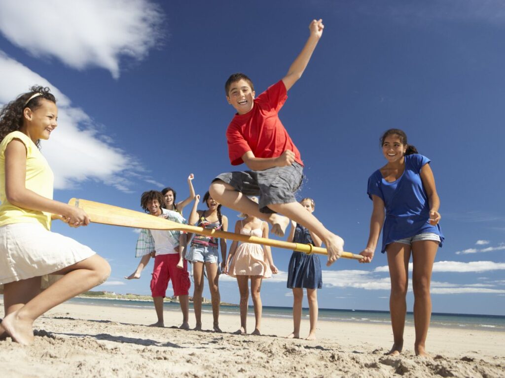 Group of joyful teenagers playing at the beach, one boy mid-air jumping over a bat held by two friends