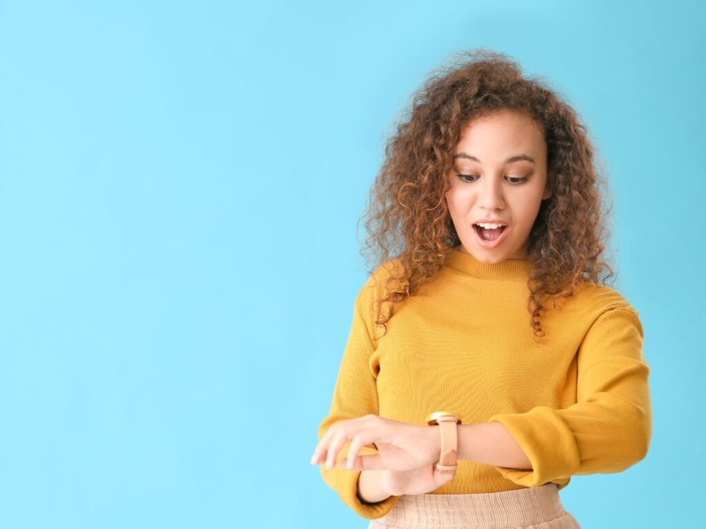 A surprised woman with curly hair and a mustard sweater looks at her smartwatch against a blue background