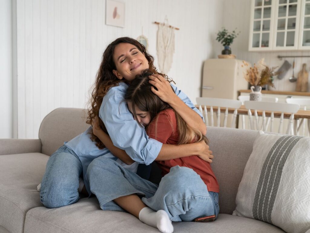A woman and a teen girl embrace warmly on a cozy sofa in a well-lit, modern living room.