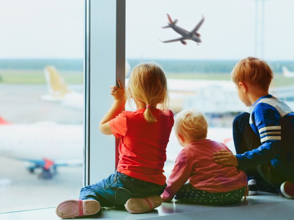 Three children sit at an airport window, watching planes. A young girl in red reaches towards a plane, while an older boy gently holds a toddler.