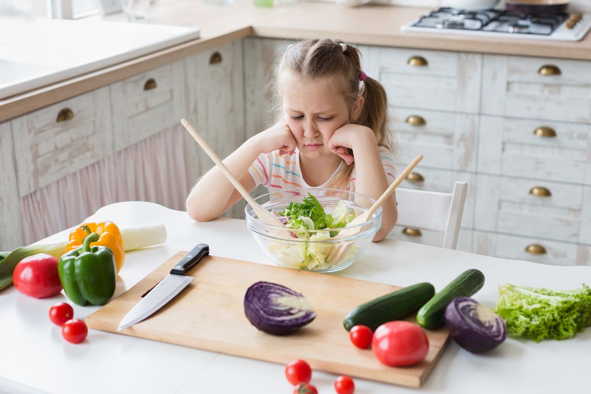 Young girl sitting at a kitchen table looking unhappy with a bowl of salad and fresh vegetables around her.