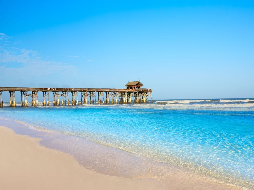 A picturesque beach scene with turquoise waters and gentle waves. A rustic wooden pier extends into the ocean under a clear blue sky