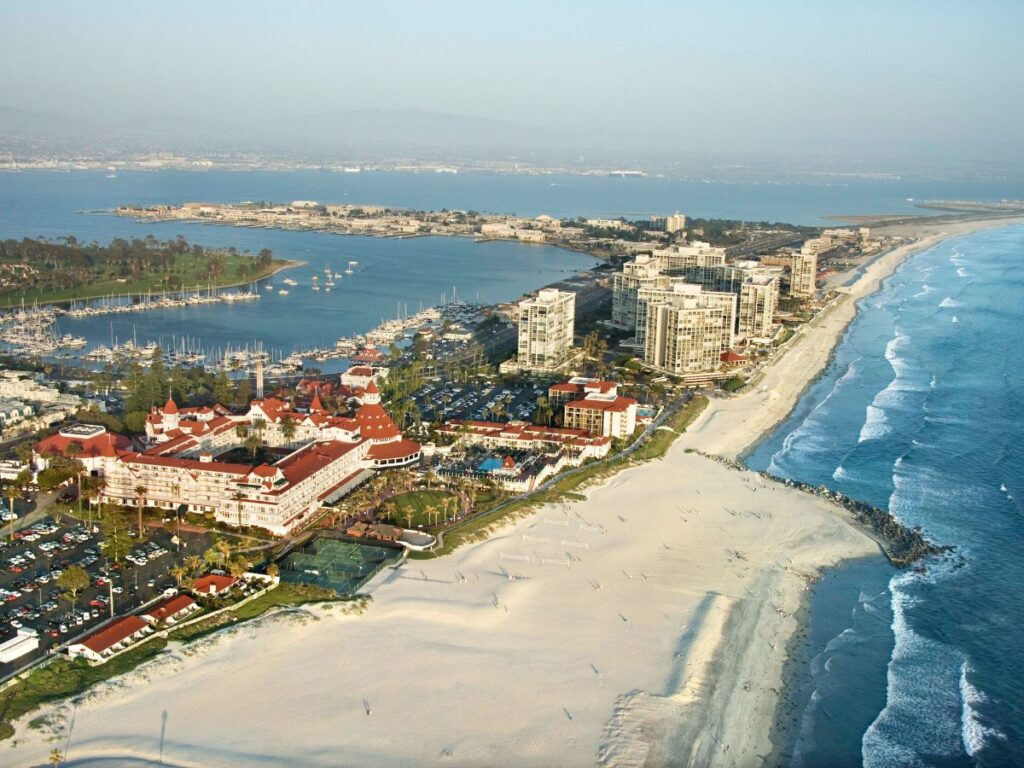 Aerial view of the coastal city and large Coronado beach, resort with red roofs, high-rise buildings, marina, and calm blue ocean