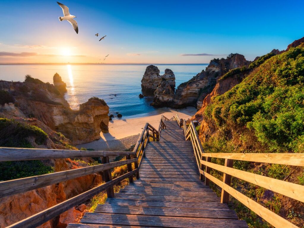 A wooden staircase leads down to a serene beach at sunrise in Algarve, Portugal, with seagulls flying and rocky cliffs framing the view