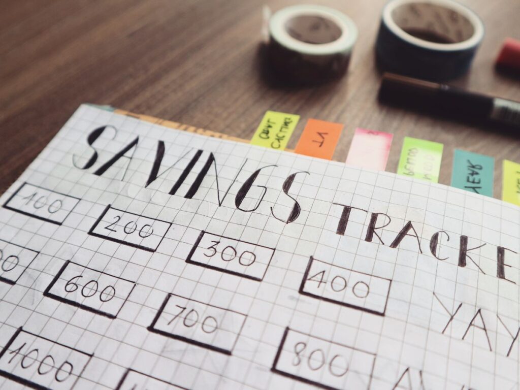 A savings tracker on graph paper with handwritten numbers in boxes, including 100, 200, and 300. Colorful sticky notes and tapes on a wooden desk.