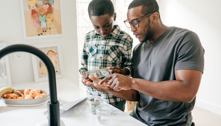 A father and son stand in a bright kitchen counting money. The father, wearing glasses, explains something to his son, who is holding dollar bills.